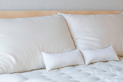 A neatly made bed features a tufted white mattress and smooth white pillows, including Shepherd's Dream Neck Pillow. The light wood headboard complements the two large pillows and two smaller ones, all in a uniform white color, creating a clean and minimalist look.