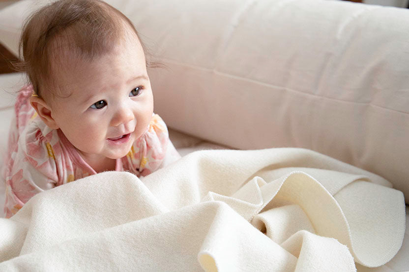A baby in a pink patterned outfit is lying on a soft, cream-colored blanket next to the Wool Filled Body Pillow by Shepherd's Dream. The baby gazes upward with a curious expression, gently lit by soft natural lighting.