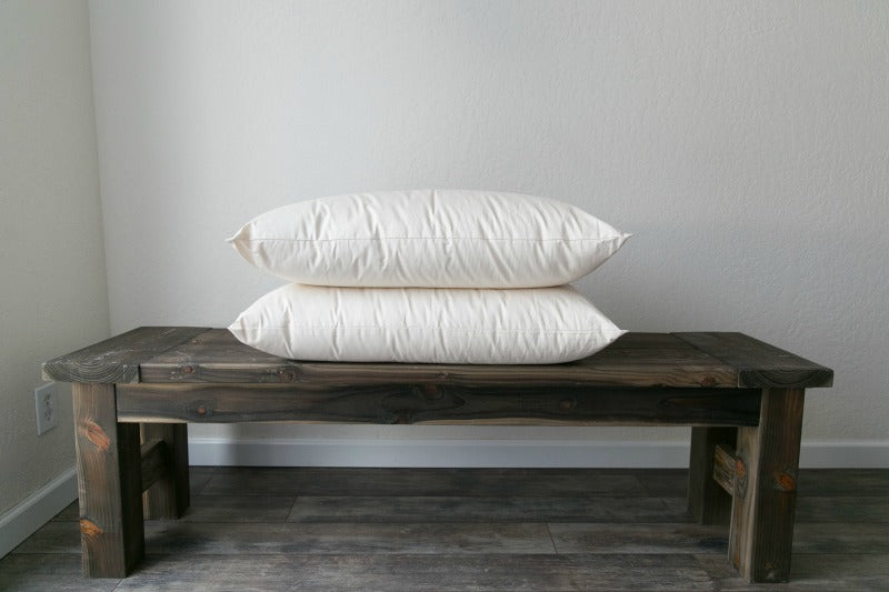 A wooden bench against a white wall features two large Shepherd's Dream Ashland Organic Wool Pillows stacked on top. The bench rests on a gray, textured floor, adding a touch of elegance to the minimalist setting.