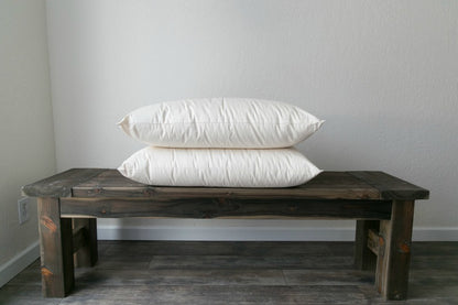 A wooden bench against a white wall features two large Shepherd's Dream Ashland Organic Wool Pillows stacked on top. The bench rests on a gray, textured floor, adding a touch of elegance to the minimalist setting.