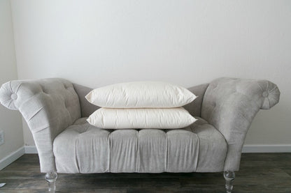 A small, gray tufted sofa with transparent legs is placed against a plain white wall. Two stacked Ashland Organic Wool Pillows by Shepherd's Dream are centered on the sofa, while the hardwood floor beneath it remains visible.