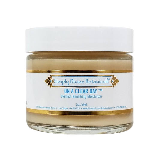 A jar of Simply Divine Botanicals On A Clear Day Blemish Banishing Moisturizer with vegan ingredients, a white lid, and a gold-accented label featuring blue text on a white background.