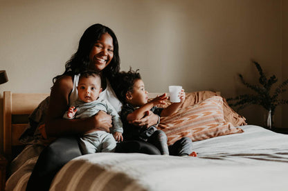 A woman is seated on a bed, smiling as she cradles a baby on her lap. Beside her, a toddler holds a white cup. The room's neutral tones are beautifully complemented by the Wool Filled Body Pillow from Shepherd's Dream, featuring EcoWool® fill. Soft pillows and an elegant plant complete the serene setting in the background.
