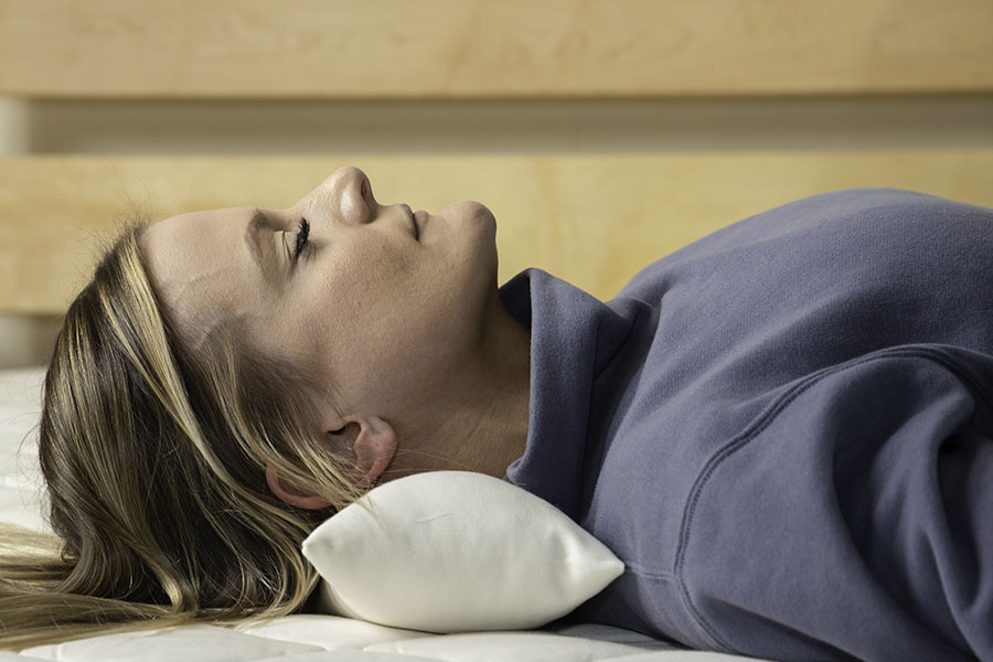 A person lies on their back on a mattress, using a Shepherd's Dream Neck Pillow for support under their neck. They are dressed in a grey sweatshirt, with the backdrop showcasing a light wooden headboard.