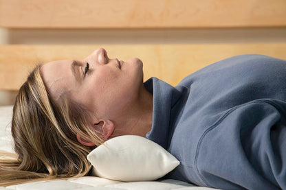 A person lies on their back on a mattress, using a Shepherd's Dream Neck Pillow for support under their neck. They are dressed in a grey sweatshirt, with the backdrop showcasing a light wooden headboard.