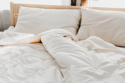 A cozy unmade bed with soft white linens, two large pillows, and sunlight streaming through a window features the Ashland Organic Wool Comforter by Shepherd's Dream. The comforter has a slightly ruffled look that makes it appear comfortable and inviting.