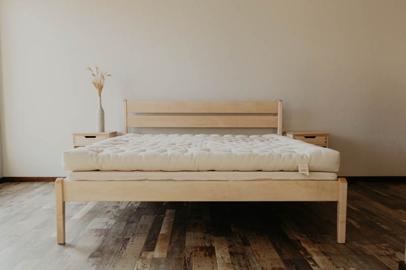 A minimalist bedroom showcasing a wooden bed frame with a white, tufted mattress resting on the Cascade Latex Mattress Base by Shepherd's Dream. Two matching wooden nightstands are placed on either side of the bed, each decorated with a vase and dried flowers. The room features hardwood flooring, and the walls are painted in a soft beige tone.