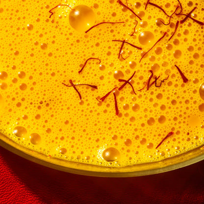A close-up of a frothy caffeine-free saffron latte, showcasing visible bubbles and delicate strands of saffron on top. The latte, made from THE FULLEST’s Warm Feelings 100-Day Supply, is served in a bowl set against a red surface. This vibrant interplay of yellow and red enhances mood improvement with every soothing sip.