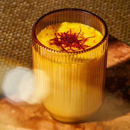 A glass from THE FULLEST's Warm Feelings 100-Day Supply is filled with creamy mango lassi, topped with a therapeutic dose of saffron. The glass features vertical grooves and sits on a textured surface under warm lighting.