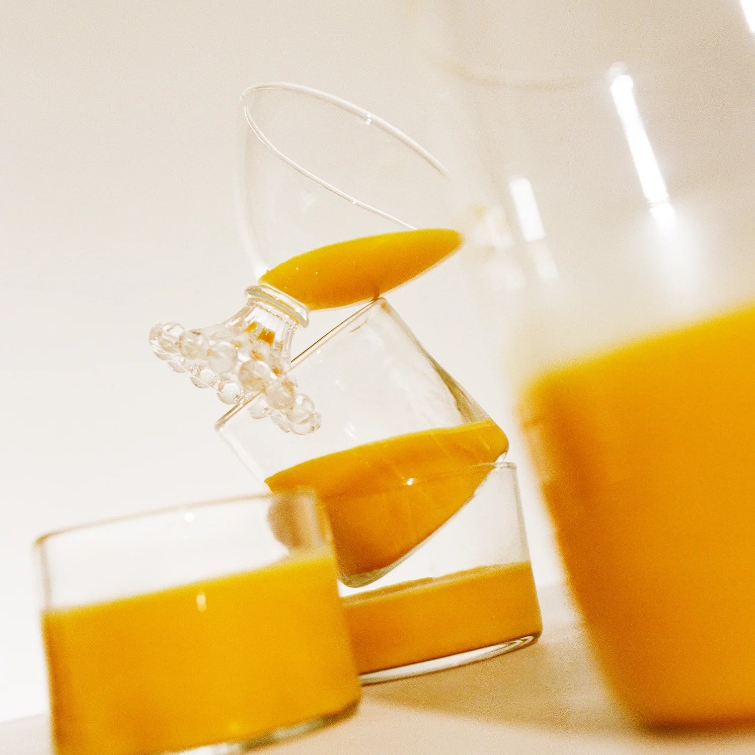 In a creative display similar to THE FULLEST's Warm Feelings 100-Day Supply, three glasses are balanced skillfully with mango juice. Two are tilted with one glass resting inside the other, and a third glass is positioned in the foreground, all set against a simple off-white background.