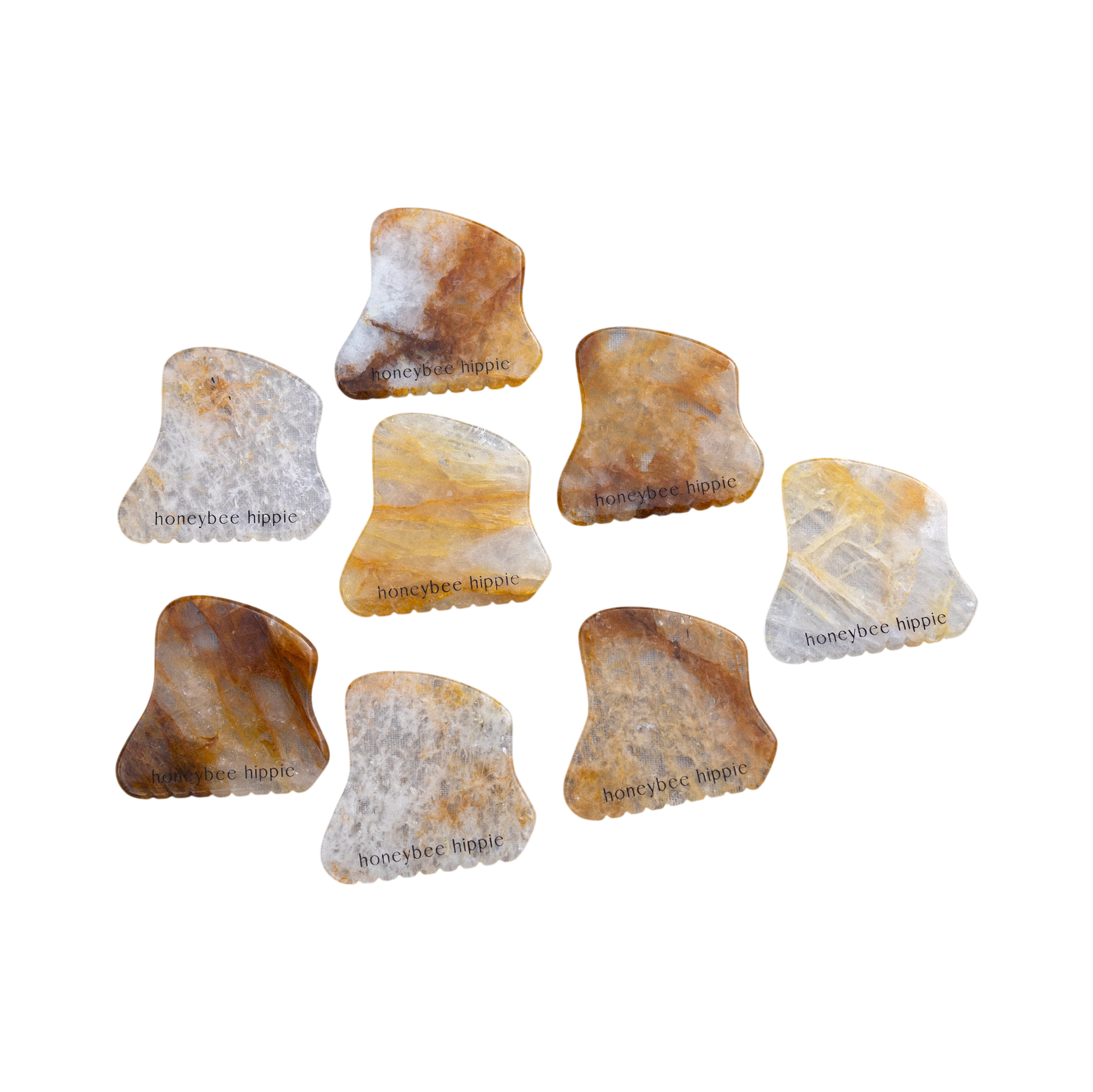 Eight Honeybee Hippie golden quartz gua sha tools with marbled brown, yellow, and gray tones—some engraved with "honeybee hippie"—are scattered on a white background.