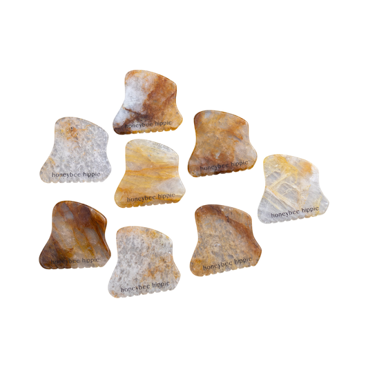 Eight Honeybee Hippie golden quartz gua sha tools with marbled brown, yellow, and gray tones—some engraved with "honeybee hippie"—are scattered on a white background.