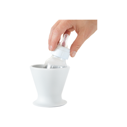 A hand smoothly performs one-handed bottle prep by placing a pink BPA-free silicone baby bottle from bökee into a stylish, white conical bökee Baby Bottle Holder, designed with a wide base. The clear bottle, featuring a white cap and nipple, fits seamlessly inside against a simple white background.