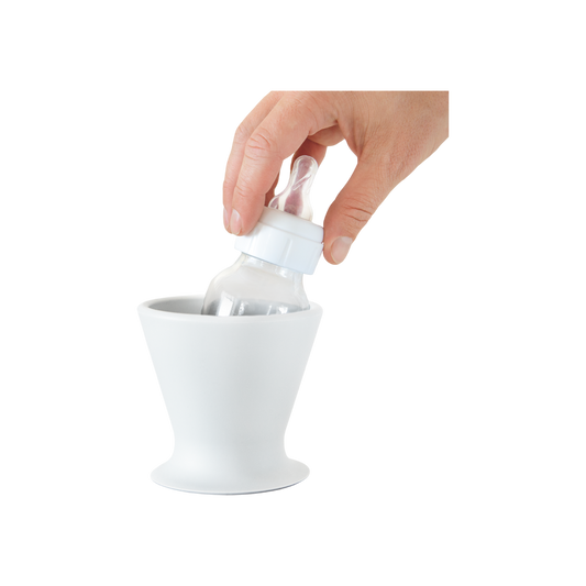 A hand smoothly performs one-handed bottle prep by placing a pink BPA-free silicone baby bottle from bökee into a stylish, white conical bökee Baby Bottle Holder, designed with a wide base. The clear bottle, featuring a white cap and nipple, fits seamlessly inside against a simple white background.