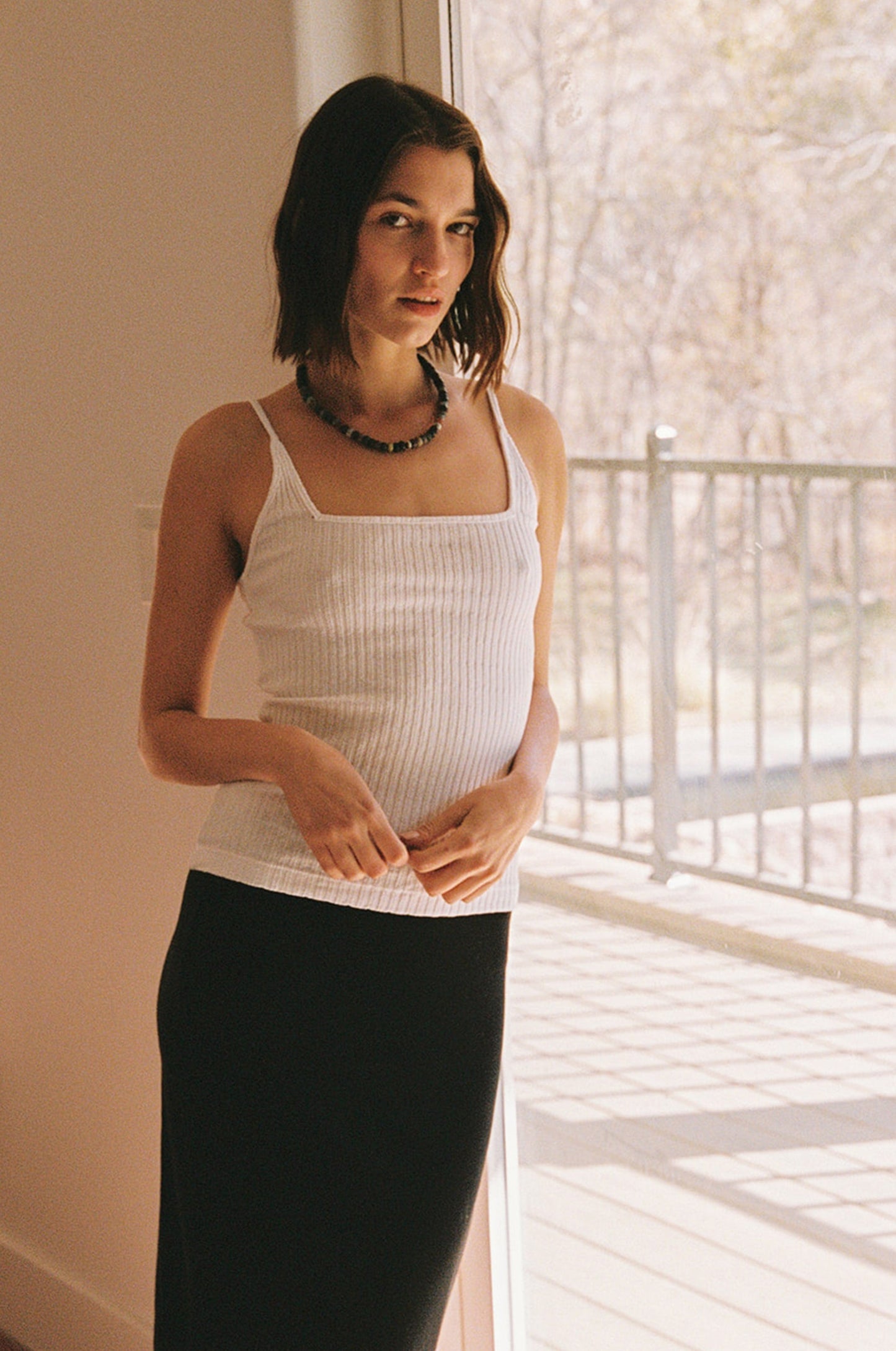 White A woman with shoulder-length brown hair wears the LA Relaxed Pointelle Square Cami, paired with a black skirt and beaded necklace, standing indoors by a sunlit glass door with an outdoor railing visible.