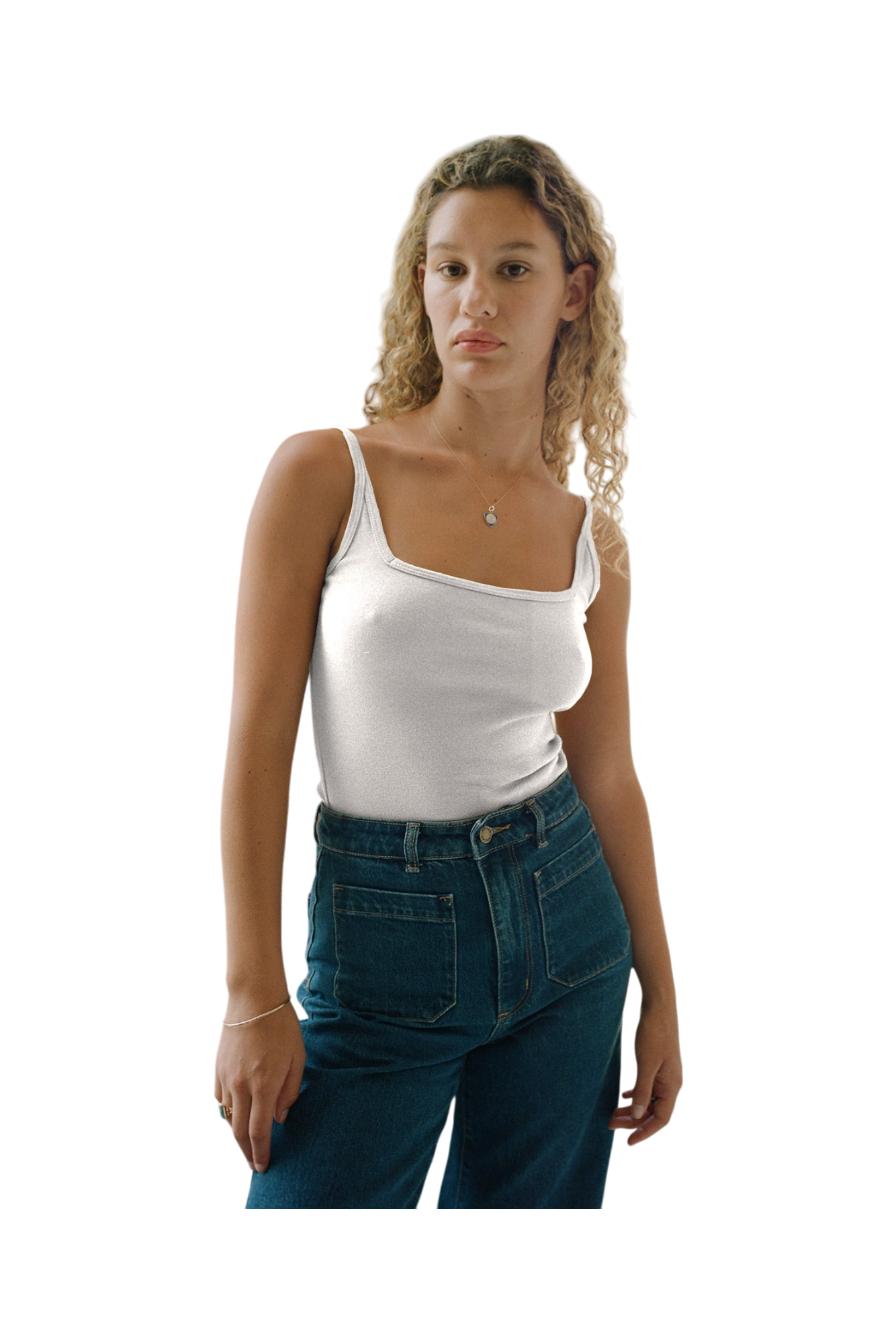 A young woman with curly blonde hair, dressed in a 90's style Organic Square Cami by LA Relaxed and high-waisted jeans, stands against a plain background.