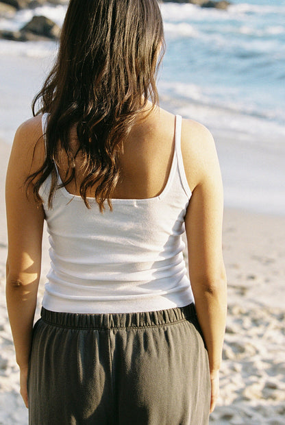 A woman with long, wavy hair stands on a sunny beach near Los Angeles, facing the ocean. She's wearing an Organic Square Cami by LA Relaxed and dark pants, with waves visible in the background.