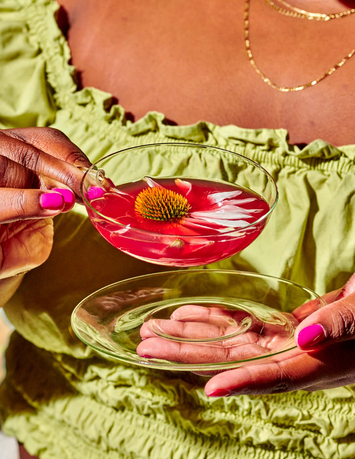 A person in a green ruffled top, with a gold necklace, holds a glass saucer and a clear glass cup filled with the pink immune toddy beverage. Inside the drink is a vibrant orange and green flower, highlighting the Lemon-Ginger Immune Toddy Sachets by WOODEN SPOON HERBS. Their nails are painted pink.
