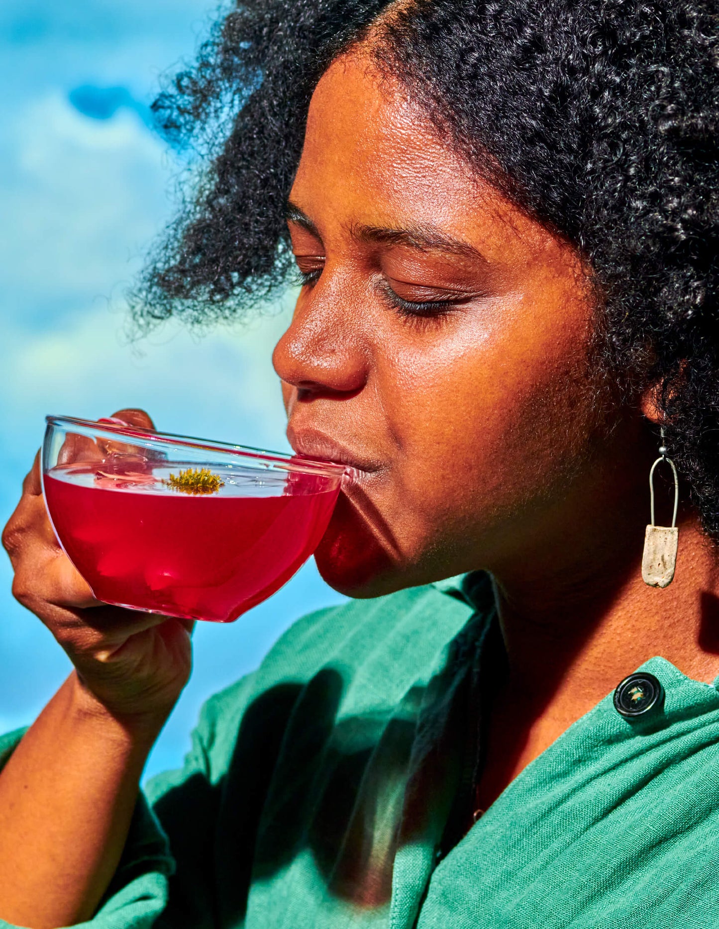 A person with curly hair sips from a clear glass teacup containing red tea. They wear a green shirt and a dangling earring, eyes closed in enjoyment. Beside them lies a jar labeled WOODEN SPOON HERBS, hinting at the Lemon-Ginger Immune Toddy Sachets inside. The background is blurred, with hints of blue and white, suggesting serenity.