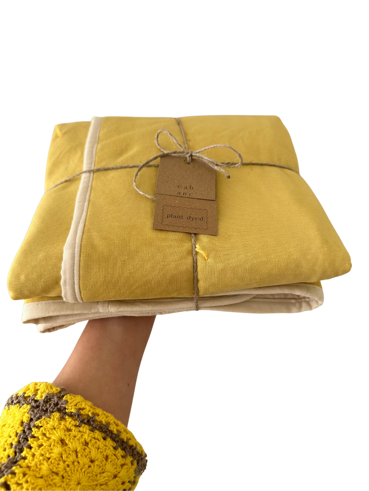 A hand in a yellow crocheted sleeve holds a folded cabane childrenswear pre-order BABY BLANKET in camomile, tied with twine and tagged “plant dyed.”.
