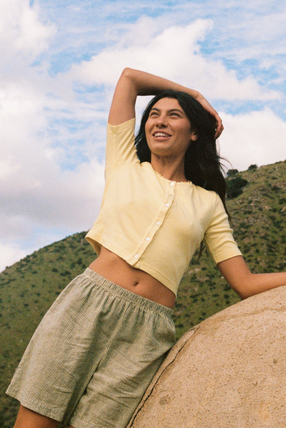 A person with long dark hair stands outdoors, smiling and leaning against a rock. They wear a Short Sleeve Organic Cardi from LA Relaxed paired with striped shorts. Green hills and a cloudy blue sky are visible behind them.